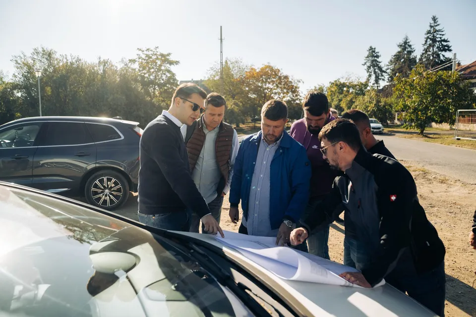 Un grup de șase bărbați stă în picioare lângă mașini și privește un document întins pe capota unei mașini albe sub lumina soarelui.