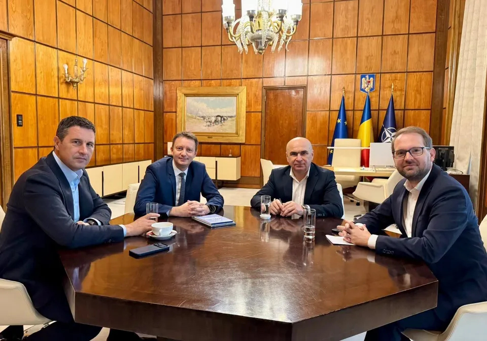 Patru bărbați în costume purtând cămăși deschise la guler stau la o masă de lemn într-o încăpere cu pereți din panouri de lemn și un candelabru.