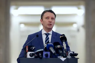 Un bărbat cu sacou albastru și cravată în dungi stă în spatele unui grup de microfoane într-o încăpere cu fundal luminos și neclar.