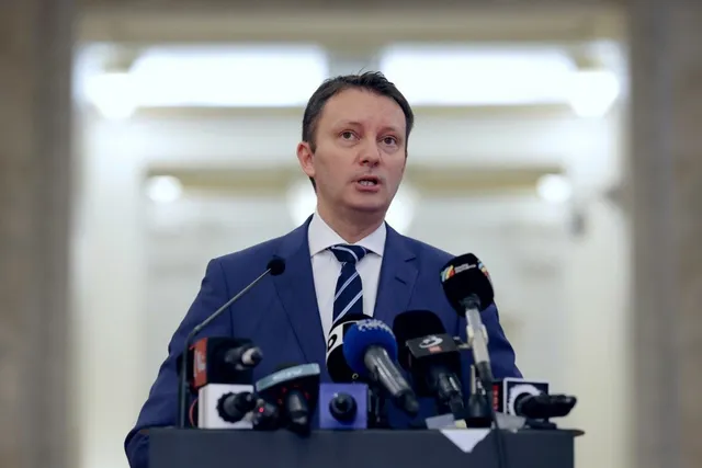 Un bărbat cu sacou albastru și cravată în dungi stă în spatele unui podium cu mai multe microfoane.