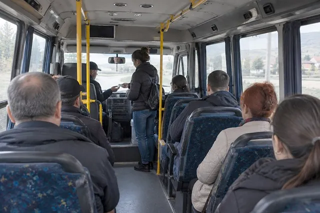 Câțiva pasageri stau pe scaunele unui autobuz, în timp ce o persoană stă în picioare pe coridor.