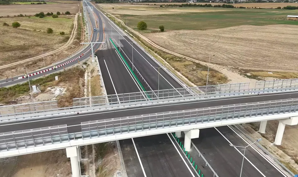 O vedere aeriană prezintă o autostradă cu mai multe benzi de asfalt, un pod peste aceasta și câmpuri deschise în fundal.