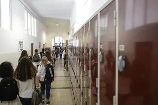 Elevi cu rucsacuri merg pe un coridor îngust lângă un șir de dulapuri metalice maronii.