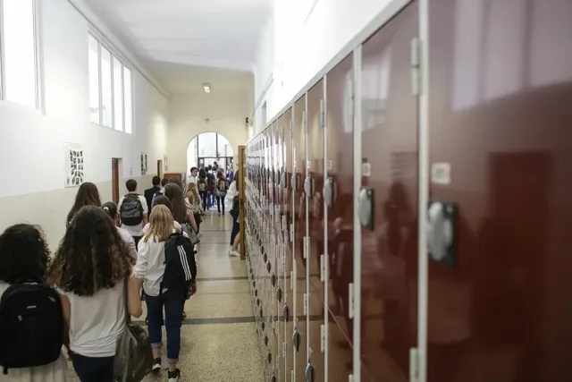 Elevi cu rucsacuri merg pe un coridor îngust lângă un șir de dulapuri metalice maronii.