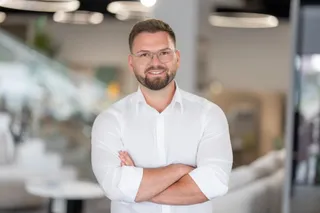 Un bărbat cu ochelari și cămașă albă stă cu brațele încrucișate într-un spațiu interior iluminat.