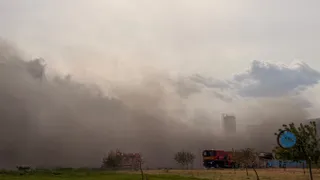 Un nor dens de fum acoperă o zonă cu clădiri și un camion de pompieri roșu parcat pe un teren deschis.