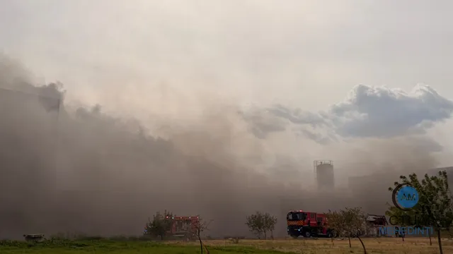 Un nor dens de fum acoperă o zonă cu clădiri și un camion de pompieri roșu parcat pe un teren deschis.