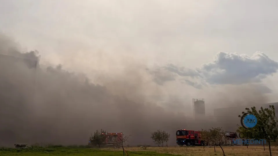 Un nor dens de fum acoperă o zonă cu clădiri și un camion de pompieri roșu parcat pe un teren deschis.