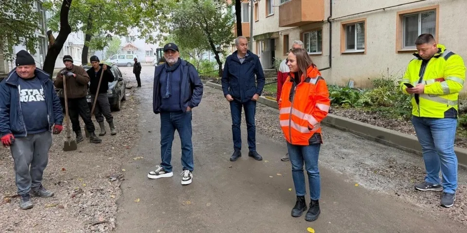 Un grup de bărbați stă pe un drum asfaltat lângă un trotuar, unii purtând haine de lucru sau jachete colorate.