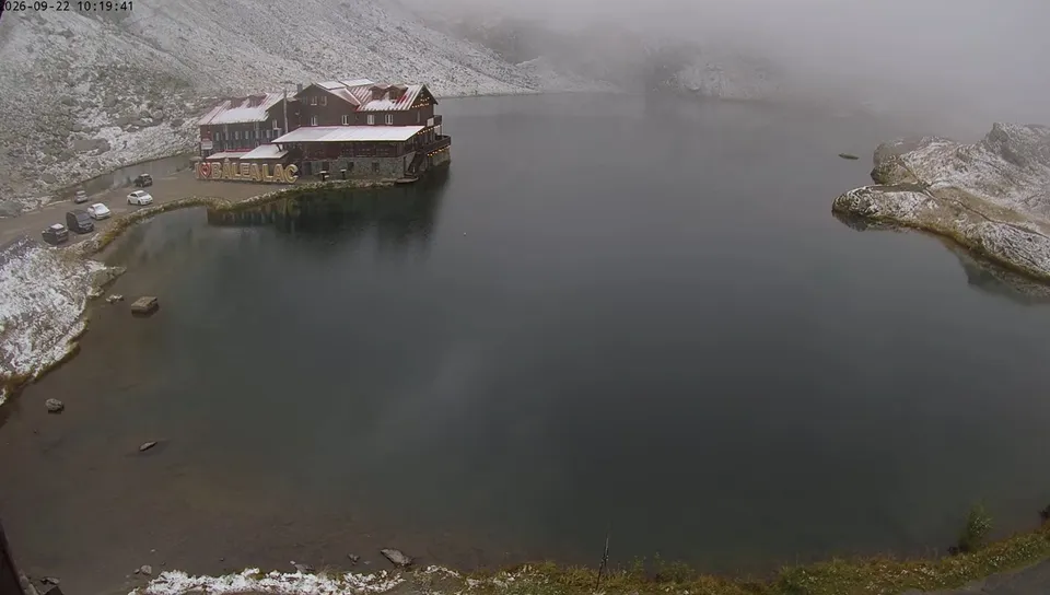 Un lac de munte este înconjurat de un teren acoperit de zăpadă și de o clădire cu acoperiș roșu sub un cer noros.