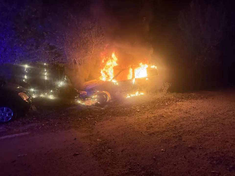 Un autoturism de culoare închisă este acoperit de flăcări pe un teren cu vegetație sub un cer nocturn.