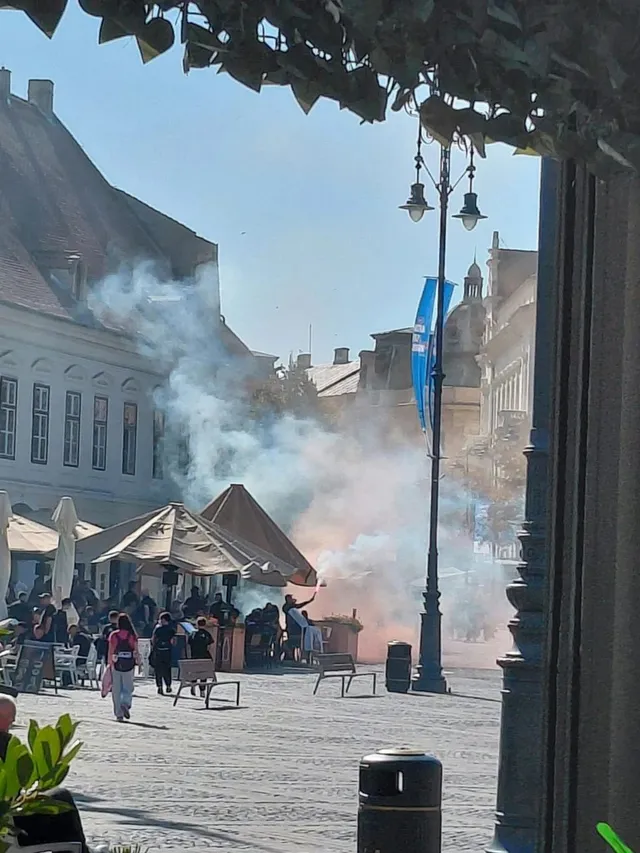 Fum alb se ridică deasupra unor umbrele de terasă și a unor persoane aflate pe o piață pietruită între clădiri urbane.