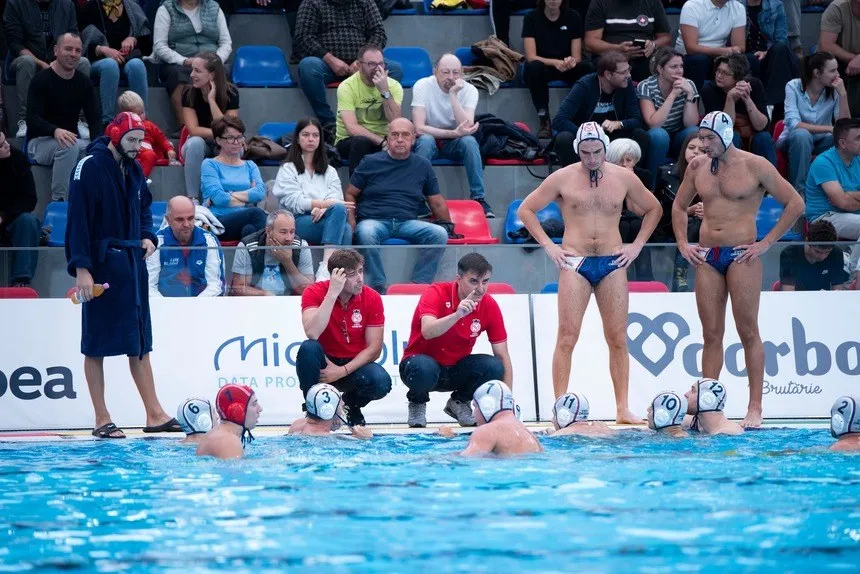 Cinci jucători de polo pe apă stau în apă, în timp ce alți doi stau în picioare lângă marginea piscinei, în fața unor spectatori așezați.