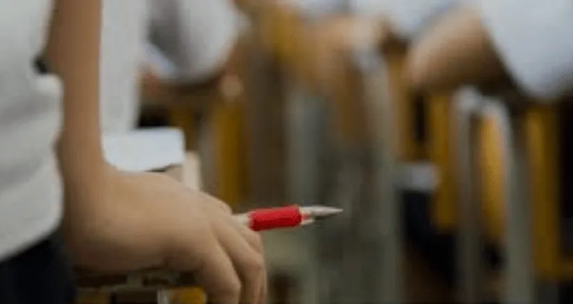 O persoană ține un pix roșu și alb în mână, fiind poziționată în fața unor obiecte de culoare galbenă pe un fundal neclar.