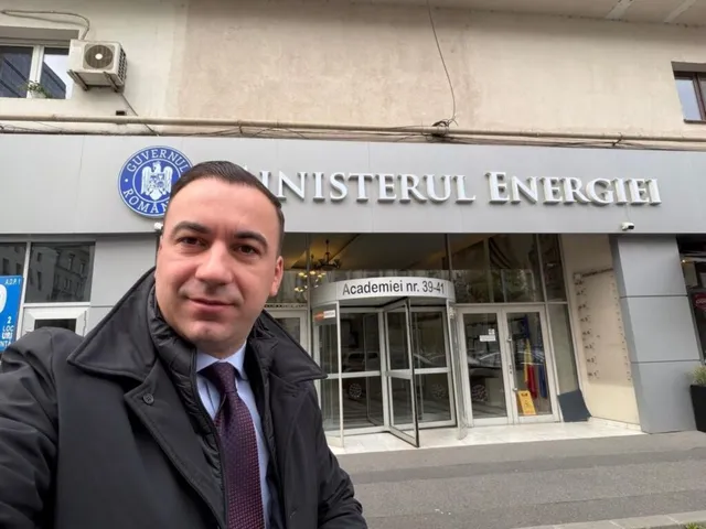 Un bărbat cu cravată și sacou negru stă în prim-plan în fața unei clădiri cu inscripția Ministerul Energiei.