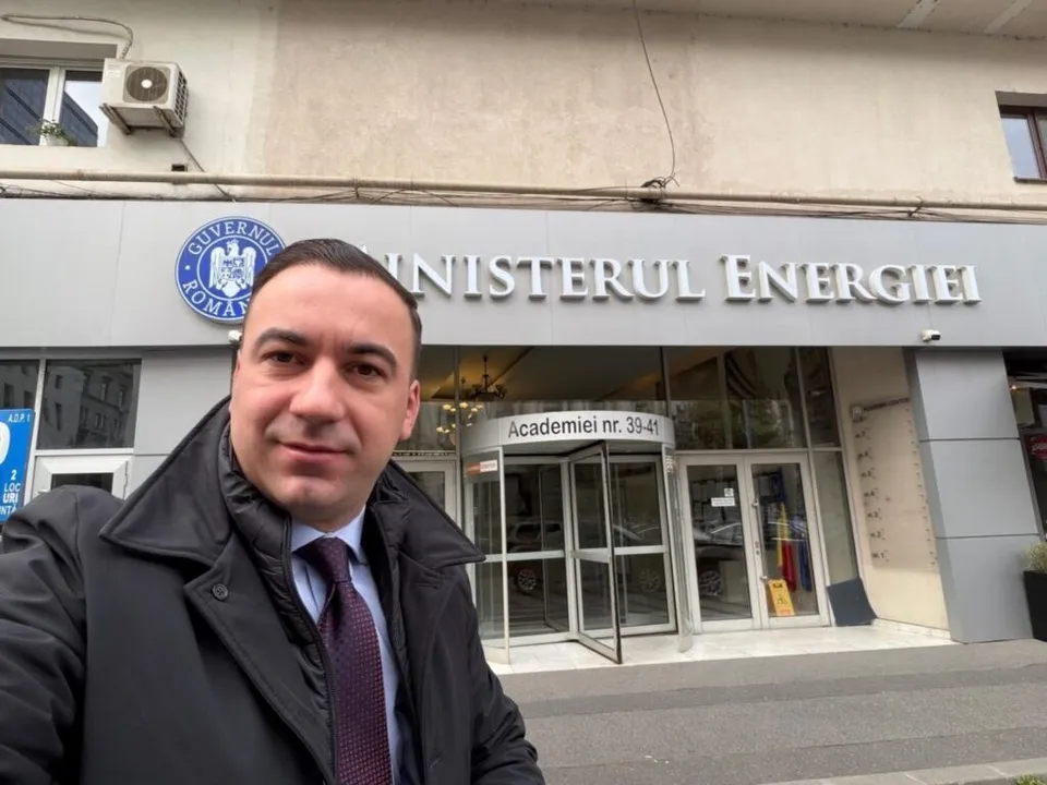 Un bărbat cu cravată și sacou negru stă în prim-plan în fața unei clădiri cu inscripția Ministerul Energiei.