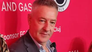 Un bărbat cu părul scurt și cămașă deschisă la guler poartă un sacou gri în fața unui fundal roșu cu text alb.