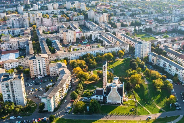 O vedere aeriană prezintă blocuri de locuințe, drumuri și un parc cu copaci verzi și o clădire cu un turn înalt.