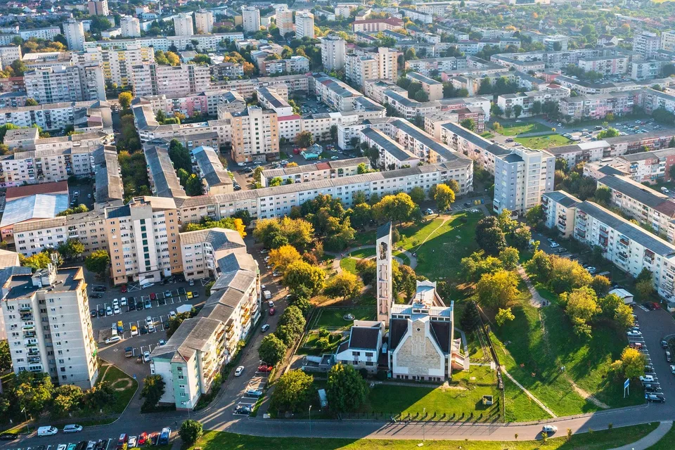 O vedere aeriană prezintă blocuri de locuințe, drumuri și un parc cu copaci verzi și o clădire cu un turn înalt.