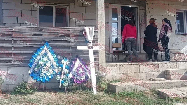 Trei coroane florale sunt așezate pe iarbă în fața unei clădiri din cărămidă, lângă o cruce albă, în timp ce trei persoane stau în prag.