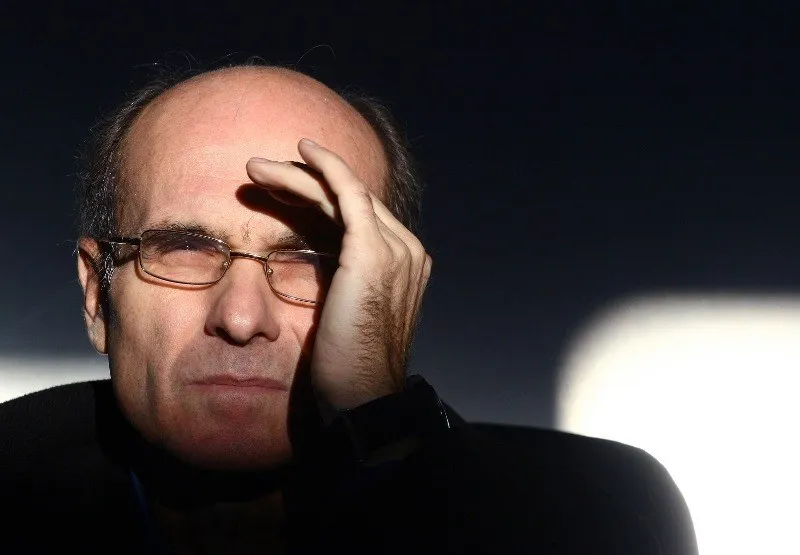 Un bărbat cu ochelari și păr rareordat își ține mâna pe frunte, privind spre lateral într-un mediu cu lumină contrastantă.