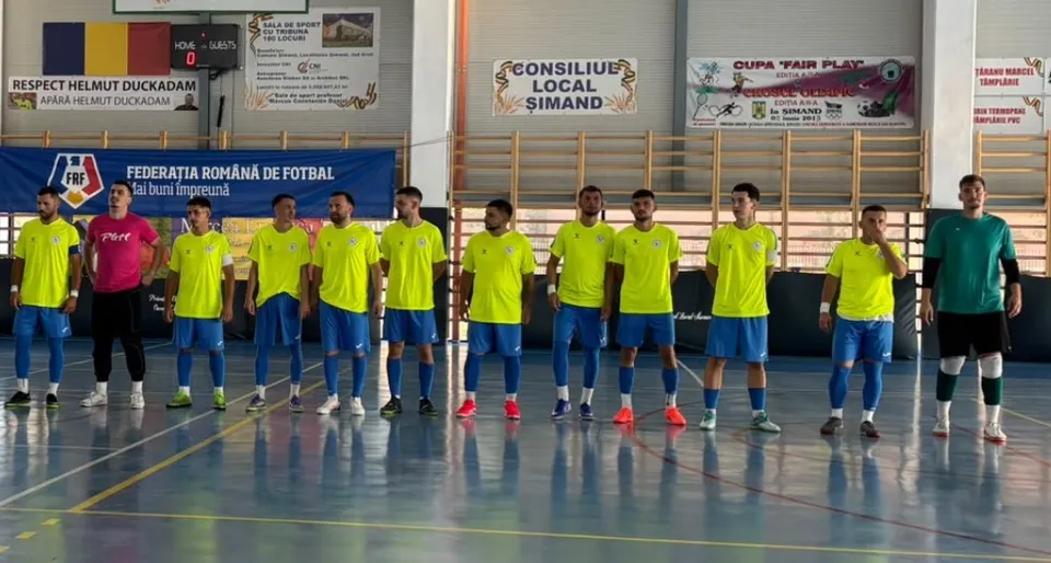 O echipă de futsal în echipamente galbene și negre stă în linie pe un teren de sport în interiorul unei săli.
