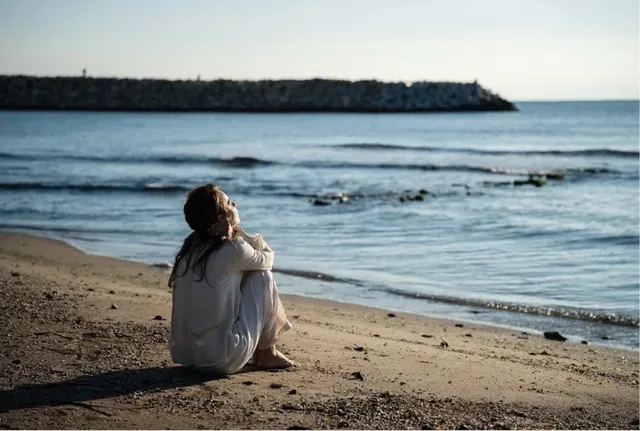 O femeie cu părul lung și haine albe stă ghemuită pe o plajă de nisip privind spre mare.