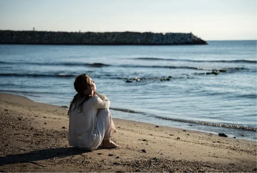 O femeie cu părul lung și haine albe stă ghemuită pe o plajă de nisip privind spre mare.