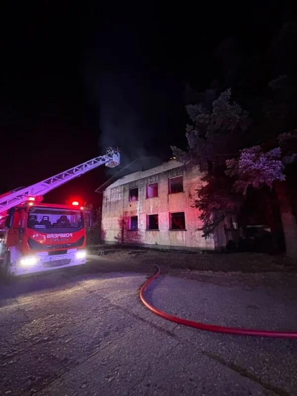 Un camion de pompieri cu o scară extinsă este poziționat lângă o clădire de două etaje în timpul nopții, cu fum vizibil deasupra acoperișului.