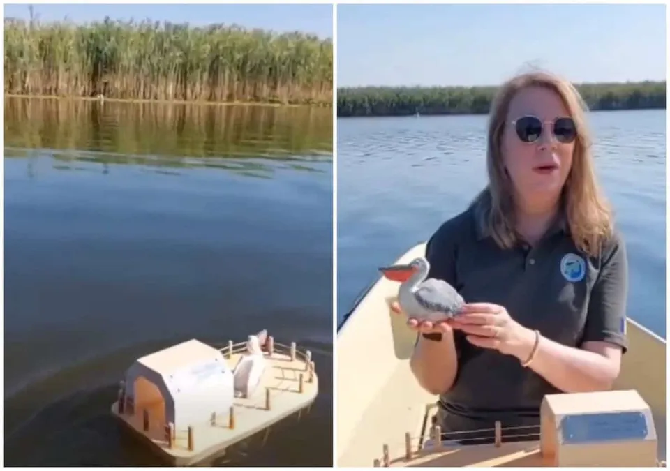 O femeie cu ochelari de soare ține o figurină de pelican pe un vas, în timp ce o mică platformă cu o clădire pluteste pe apă.