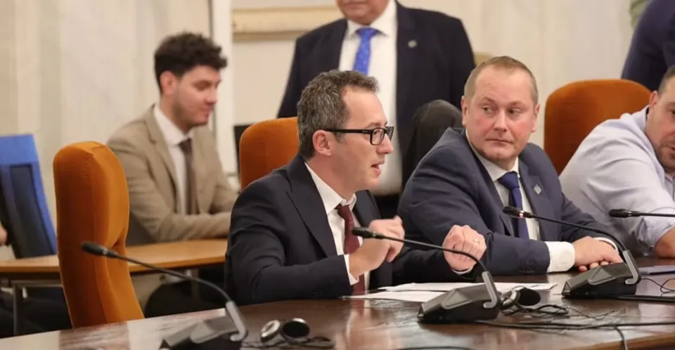 Un bărbat cu ochelari și cravată roșie vorbește la un microfon în timp ce alți bărbați stau la o masă de conferință.