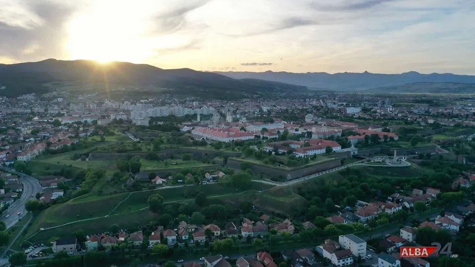 Vedere aeriană de ansamblu a unui oraș cu străzi, case, clădiri, dealuri și cer luminat de soare.