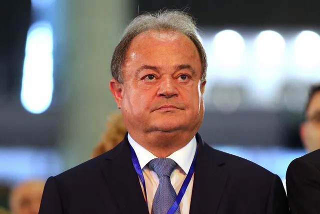 Un bărbat cu părul scurt și cărunt poartă sacou închis la culoare, cămașă albă și cravată albastru deschis cu un cordon albastru în jurul gâtului.
