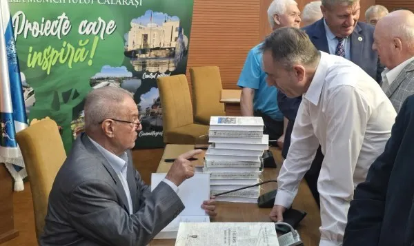 Un bărbat cu ochelari arată spre o stivă de cărți de pe o masă, în timp ce alți bărbați stau în jurul acesteia.