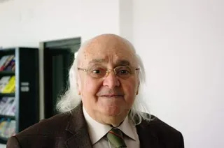 Un bărbat cu părul alb și ochelari poartă o sacoșă maro, o cămașă albă și o cravată în culori închise.