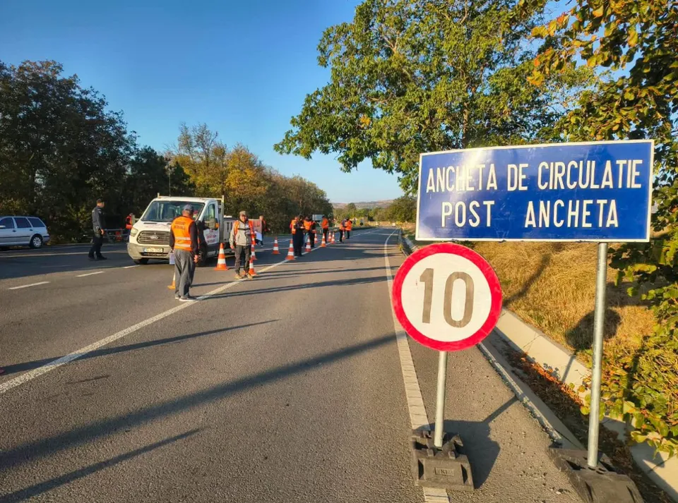 Un semn albastru cu text alb și un semn circular cu cifra zece sunt amplasate pe marginea unui drum asfaltat lângă un vehicul alb.