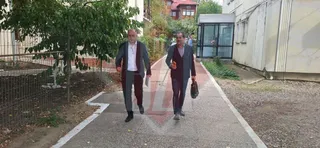 Doi bărbați merg pe o alea pavată între un gard și o clădire albă.