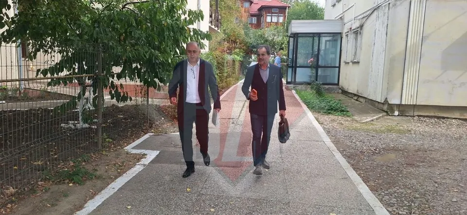 Doi bărbați merg pe o alea pavată între un gard și o clădire albă.