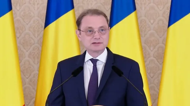 Un bărbat cu ochelari și cravată purtând un costum închis stă în fața unor steaguri galbene și albastre.