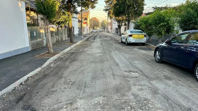 O stradă cu asfalt neuniform, borduri de beton, un trotuar, un autovehicul alb și un autovehicul negru parcate pe marginea drumului.