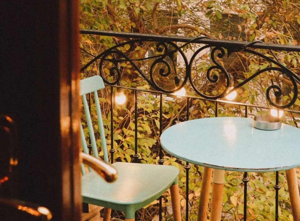 O masă rotundă și un scaun de culoare albastru deschis sunt plasate pe un balcon cu balustradă metalică decorativă și vegetație în fundal.