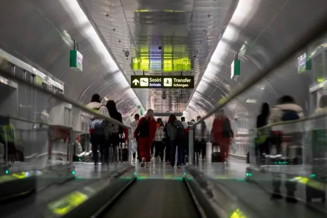 Oamenii merg pe un coridor de aeroport cu un sistem de bandă rulantă și panouri de semnalizare suspendate de tavanul metalic.