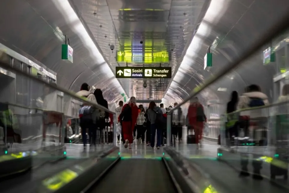 Oamenii merg pe un coridor de aeroport cu un sistem de bandă rulantă și panouri de semnalizare suspendate de tavanul metalic.