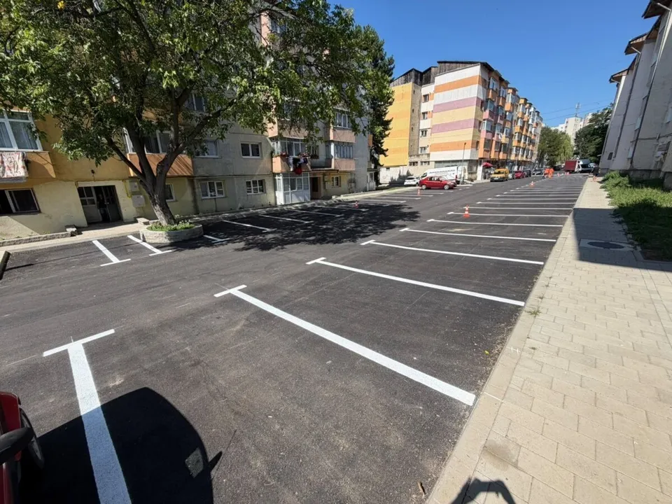 Un teren asfaltat cu linii albe pentru locuri de parcare se află în fața unor clădiri de locuințe sub un cer senin.