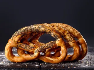 Trei covrigi de tip pretzel, unul presărat cu semințe de mac și două cu semințe de susan, sunt așezați pe o suprafață închisă.