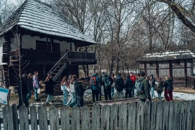 Un grup de persoane stă în fața unor construcții din lemn cu acoperișuri învelite în zăpadă, într-o zonă cu copaci fără frunze.