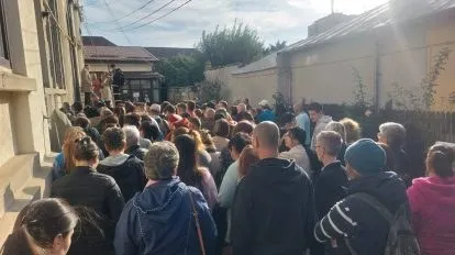 O mulțime de persoane stă în picioare pe un drum îngust între clădiri, cu spatele la cameră.