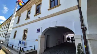 O clădire albă cu ferestre maronii dispune de un arc de intrare prin care se vede un drum și câteva siluete de persoane.