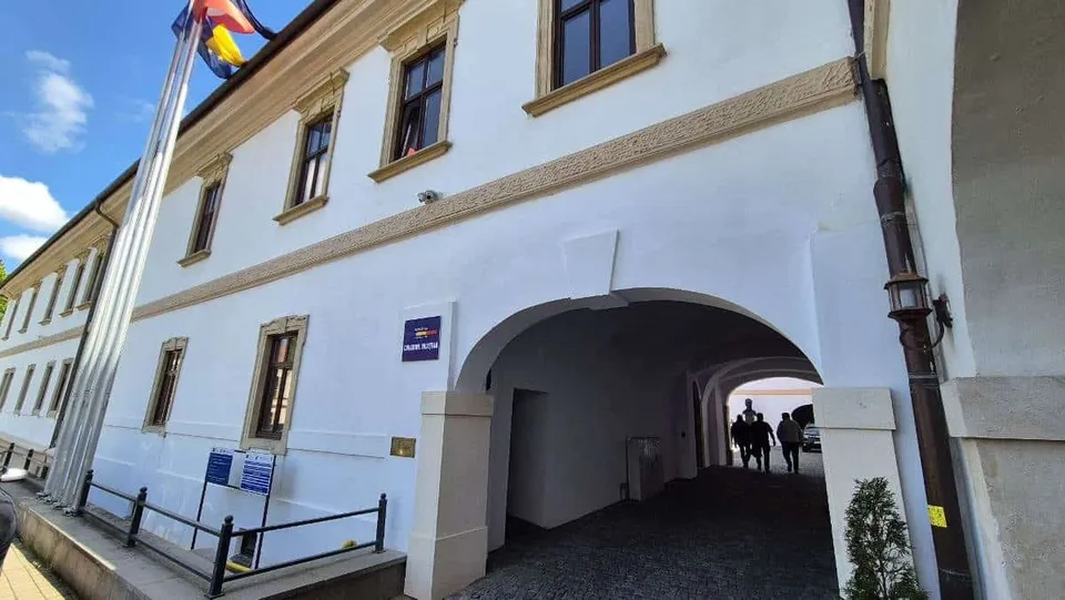 O clădire albă cu ferestre maronii dispune de un arc de intrare prin care se vede un drum și câteva siluete de persoane.