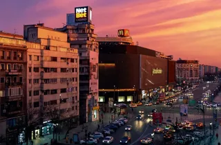O peisaj urban la apus cu clădiri, trafic intens pe șoseauă și un cer în tonuri roz și portocaliu.